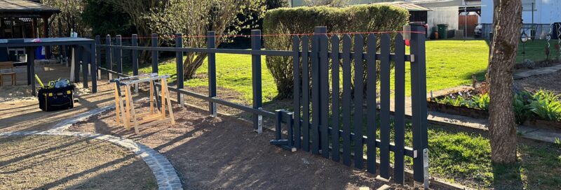 Ein individuell gefertigter Gartenzaun aus dunkel lasiertem Lärchenholz im Aufbau: Die Zaunelemente sind exakt an das unebene Gelände angepasst, mit sorgfältig gesetzten Pfosten und durchgehender Fluchtlinie. Im Bild sichtbar sind auch Bauwerkzeuge und Hilfsmittel, die auf eine saubere, handwerkliche Umsetzung hinweisen. Das Projekt liegt in einem Wohngebiet nahe Gießen, mit Blick auf angrenzende Gärten und Häuser.