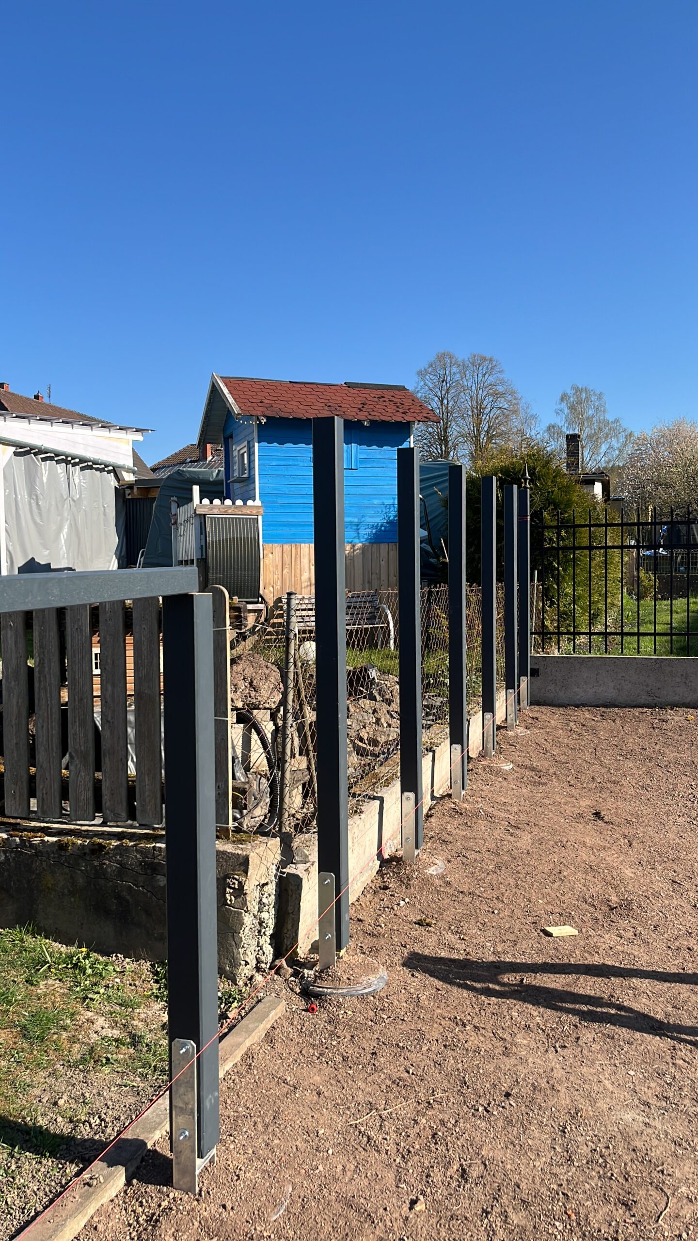 B08B21D1-B5BD-4411-B903-662BAEDA3367 Blick auf eine Baustelle mit frisch gesetzten Metallpfosten für einen Gartenzaun aus Holz. Im Hintergrund stehen ein blau gestrichenes Spielhaus und mehrere Gartenhäuser. Eine gespannte Richtschnur verläuft am Boden entlang der Zaunlinie, die sich an ein Gefälle im Gelände anpasst.