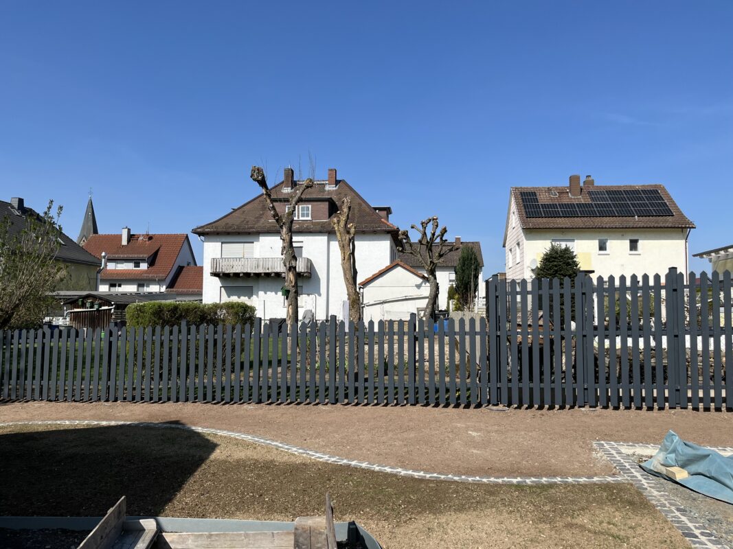 IMG_7847 Ein dunkel lasierter Gartenzaun aus Holz zieht sich entlang eines Grundstücks vor mehreren Wohnhäusern. Der Zaun ist auf der rechten Seite höher, um Sichtschutz zu bieten. Im Vordergrund sieht man einen hell gepflasterten Weg und eine Rasenfläche.